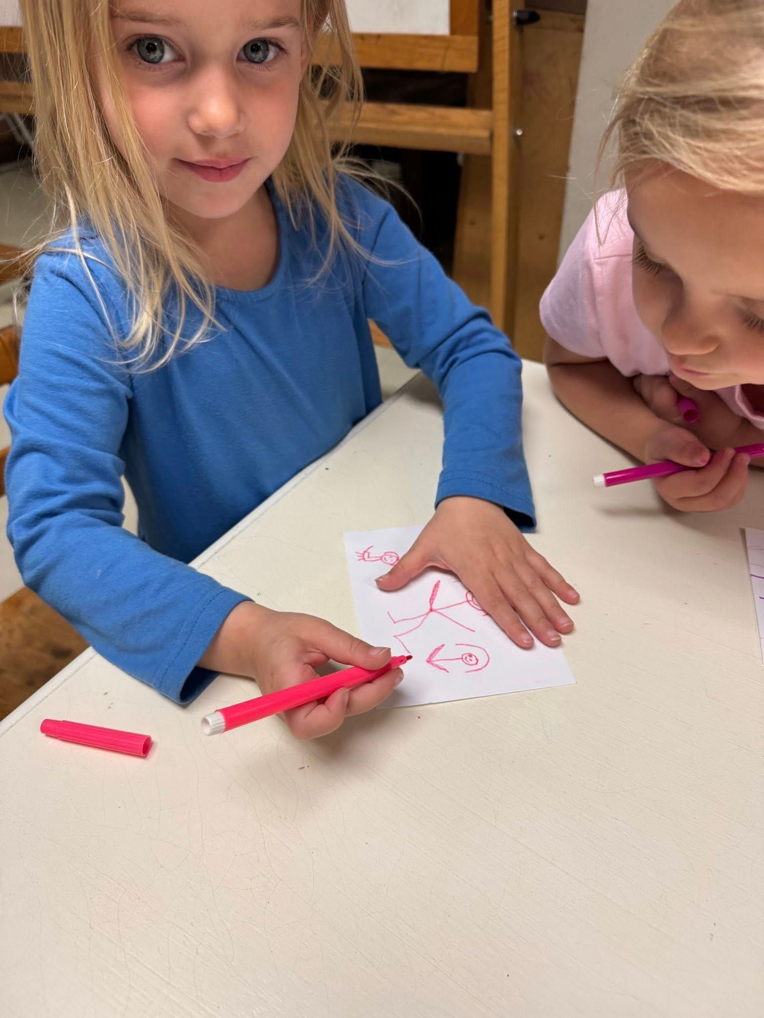 Two little girls are sitting at a table drawing with pink markers.