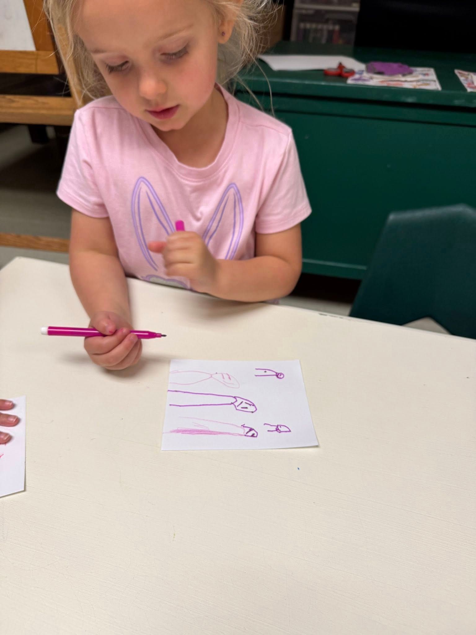 A little girl is sitting at a table holding a pink pen.