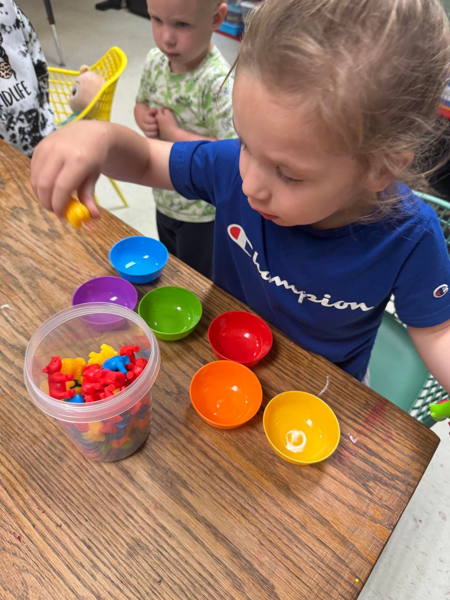 A little girl is playing with a bucket of toys on a wooden table.