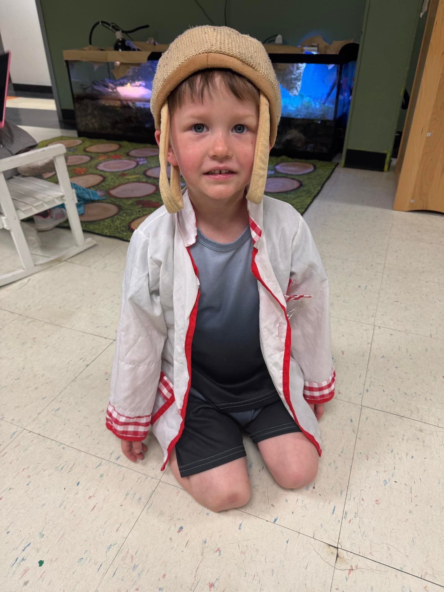 A little boy in a lab coat and hat is kneeling on the floor.