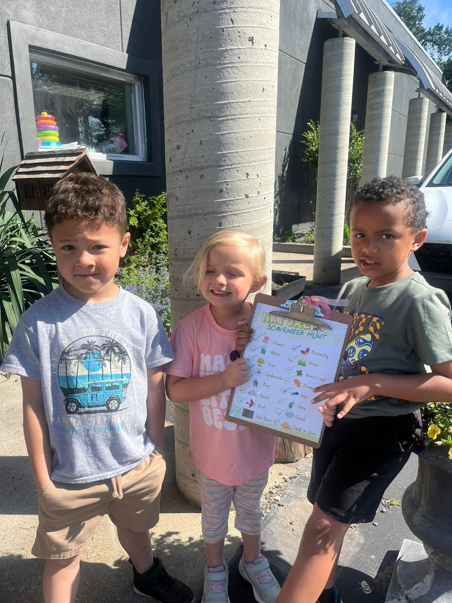 Three children are standing next to each other in front of a building holding a clipboard.