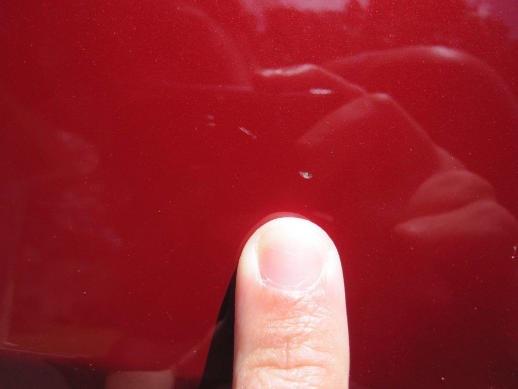 Finger pointing at a small scratch on a red car.