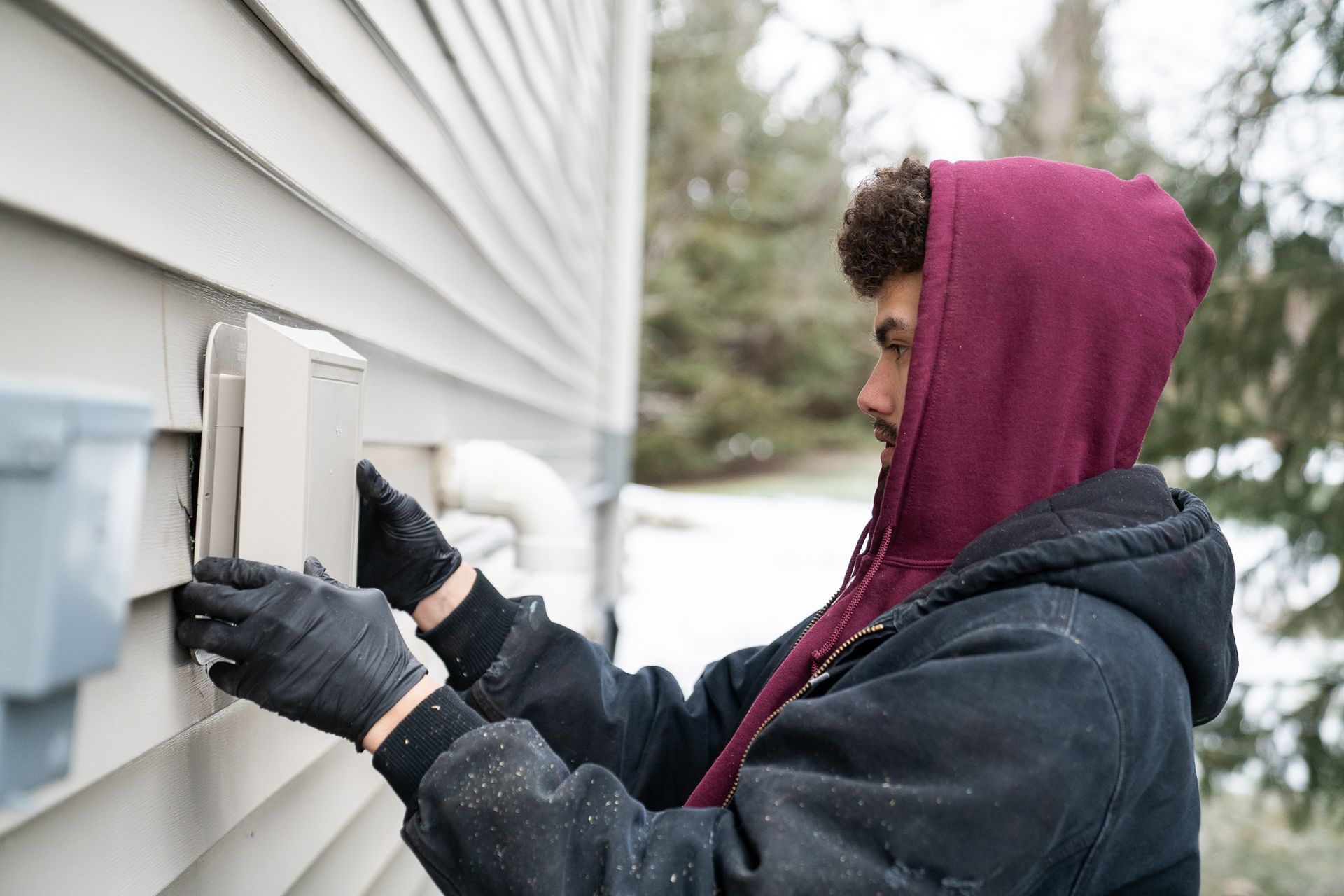 A man wearing a hoodie and gloves is installing a vent on the side of a house.