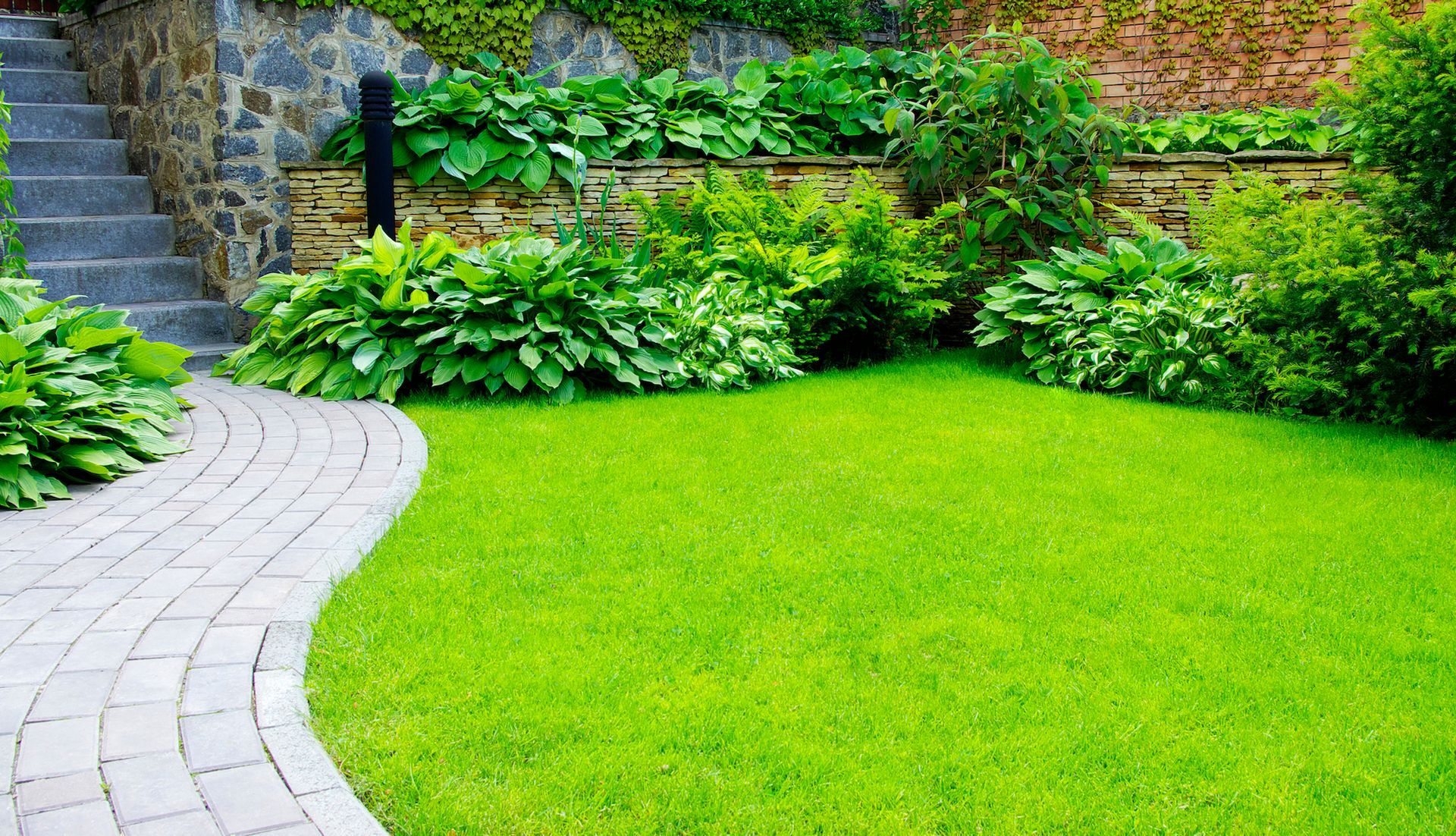 A curved stone path leads past a bright green lawn toward tiered garden beds with lush plants and a stone wall.