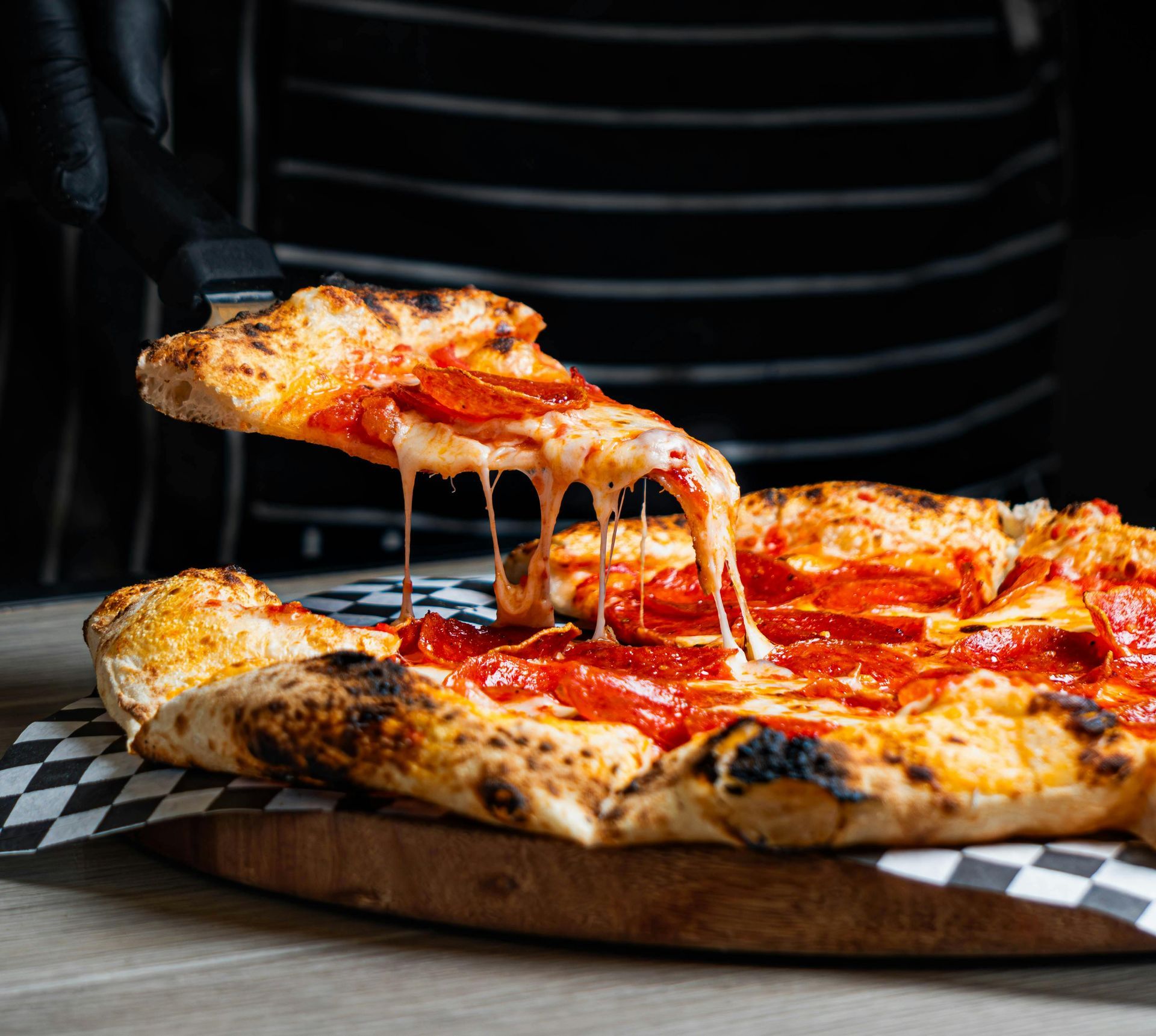 A gloved hand lifts a slice of pepperoni pizza from a wooden board, showing melted, stretching cheese.