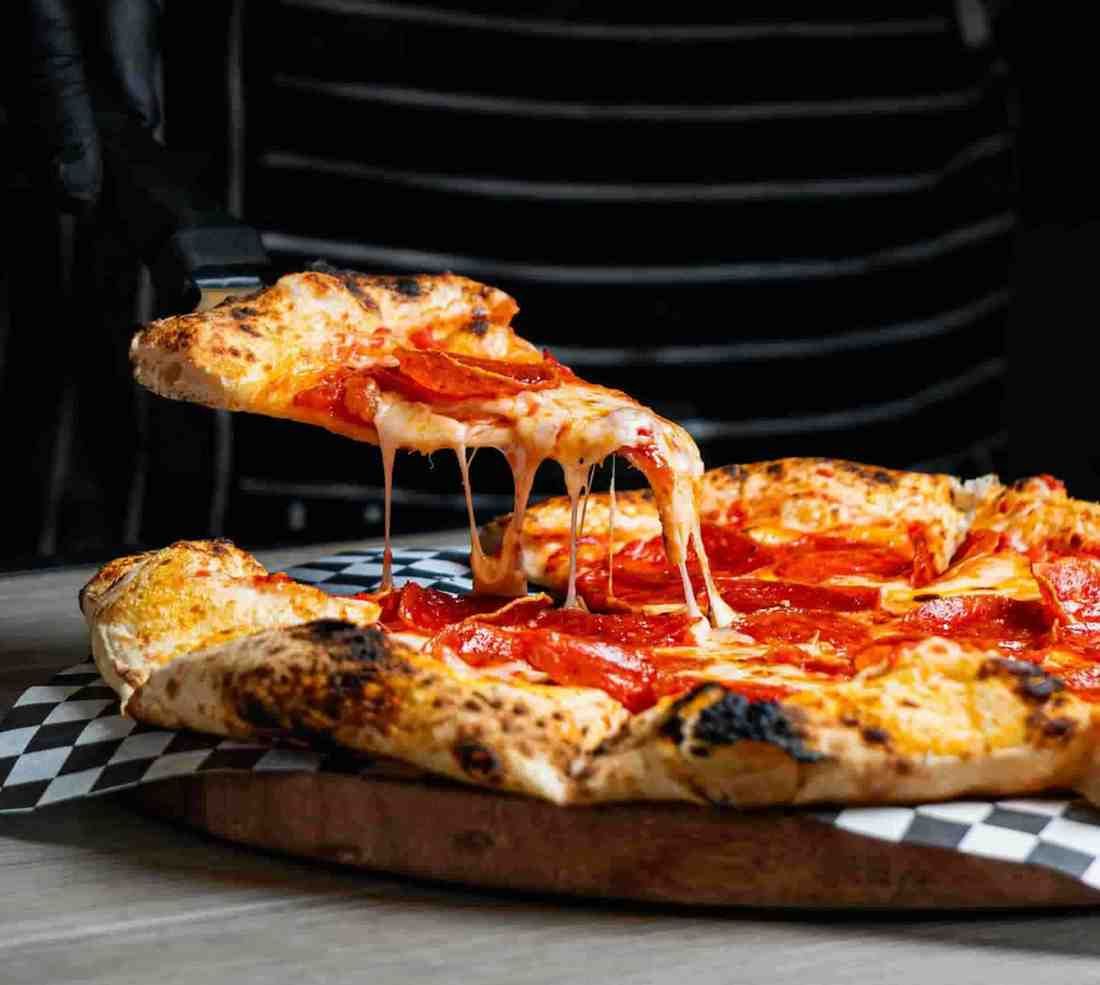 A gloved hand lifts a slice of pepperoni pizza from a wooden board, showing melted