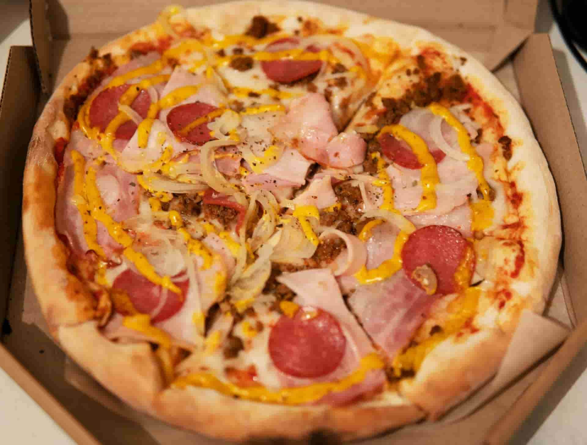 A round pizza in a cardboard box, topped with ham, pepperoni, onions
