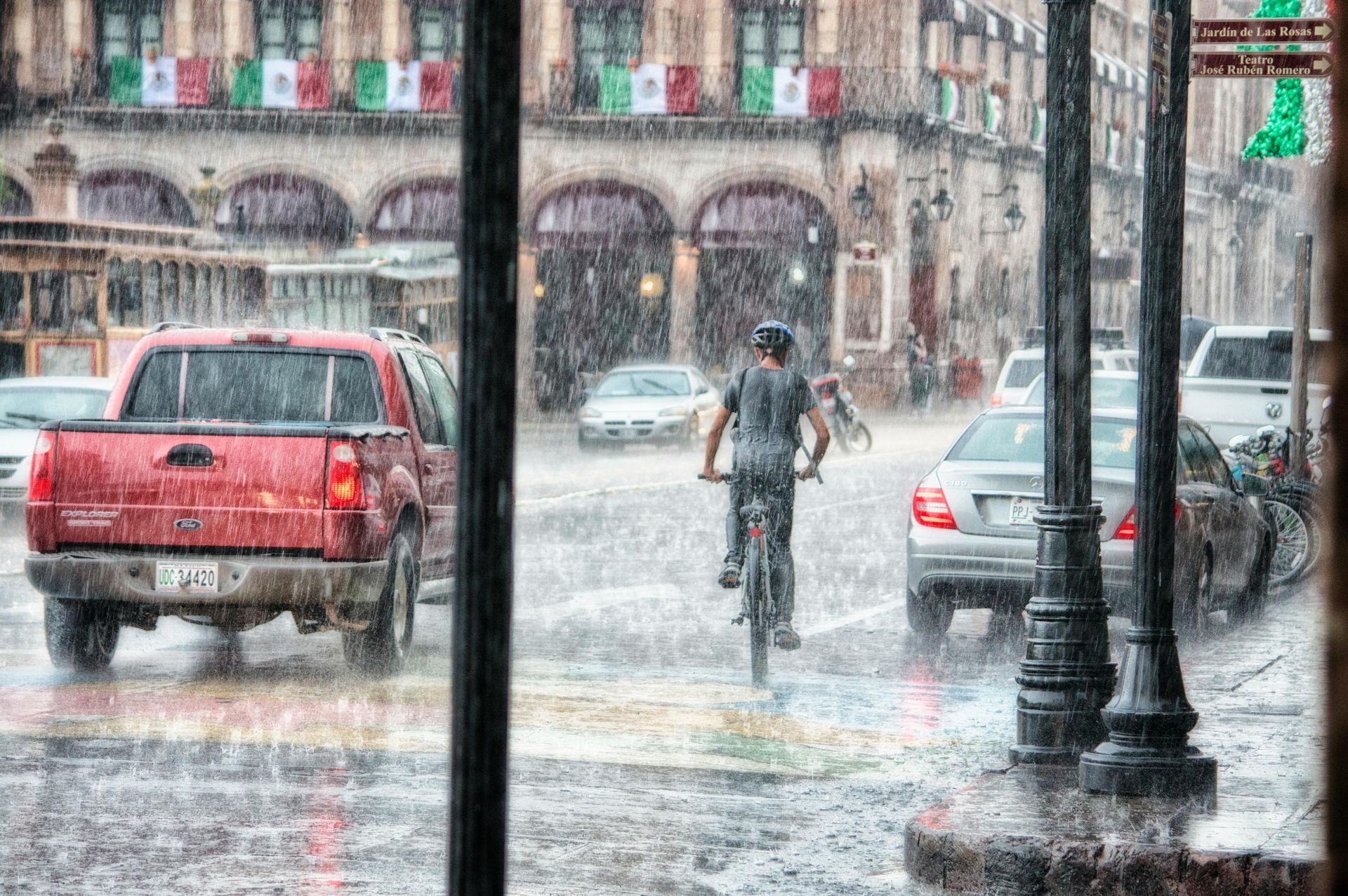 Heavy rain in a city street; a bicyclist, cars, and a red truck are visible. Mexican flags hang in the background.