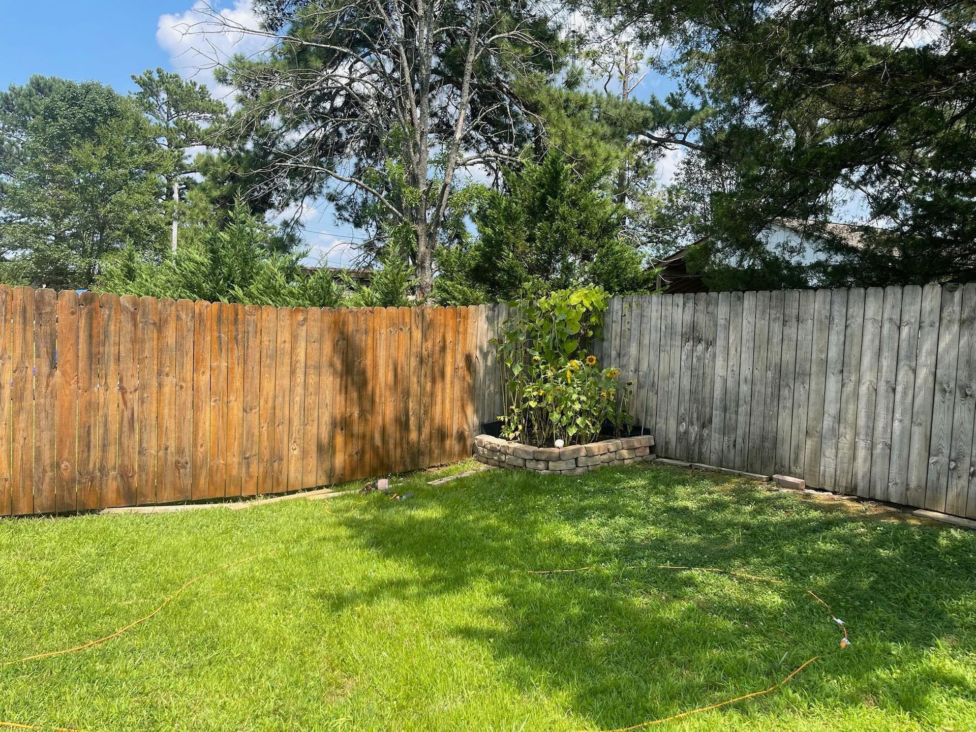 Lawn with a brown and gray wooden fence, small garden, and trees in the background on a sunny day.