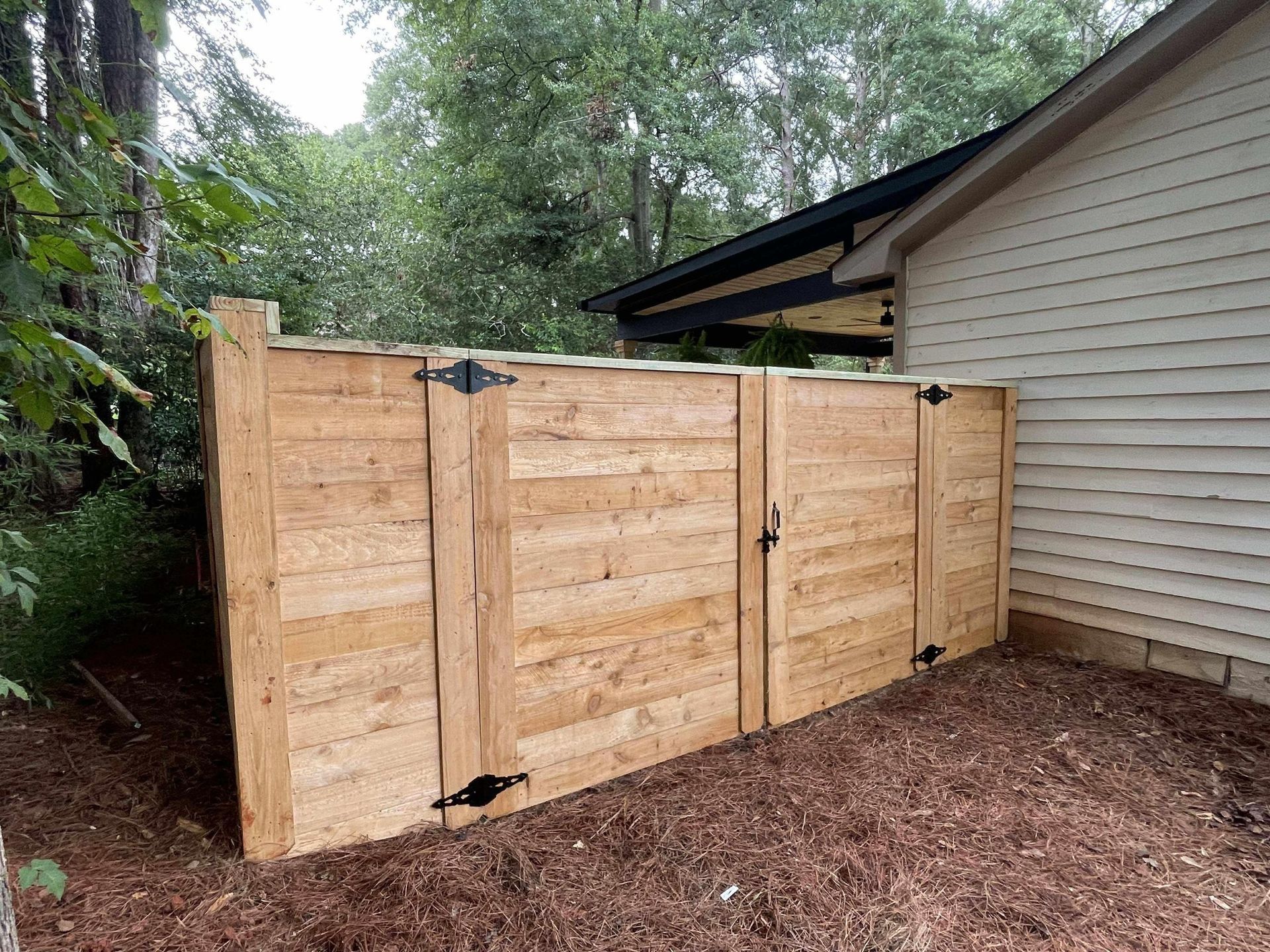 Wooden gate enclosure with black hinges and latch next to a light-colored house. Mulch ground, trees in background.