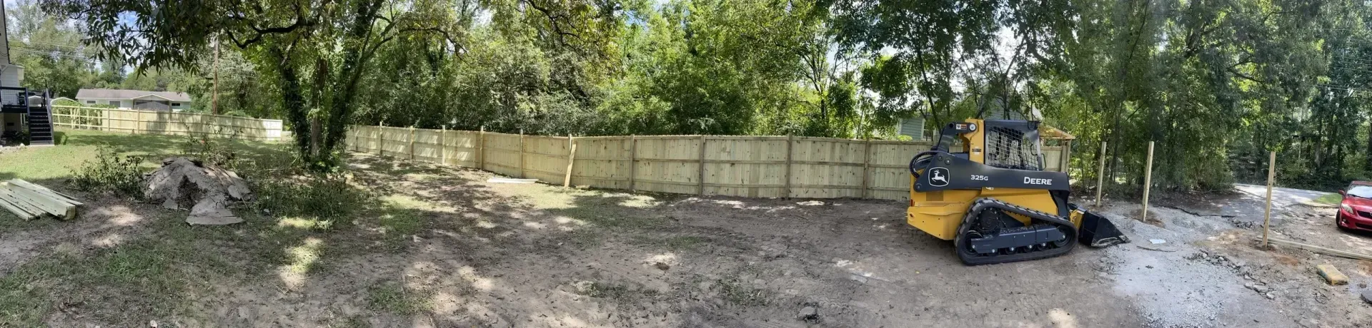 A construction site with a yellow skid steer, a fence, trees, and a red car.