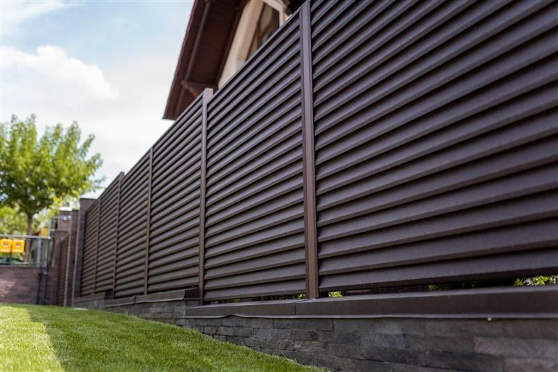 Brown slatted fence on a brick base in front of a house with green grass and a blue sky.