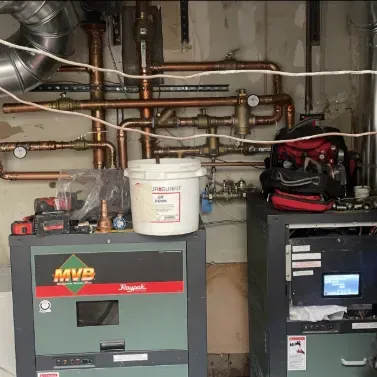 Two gas boilers with copper pipes and various tools, located in a basement or utility room.