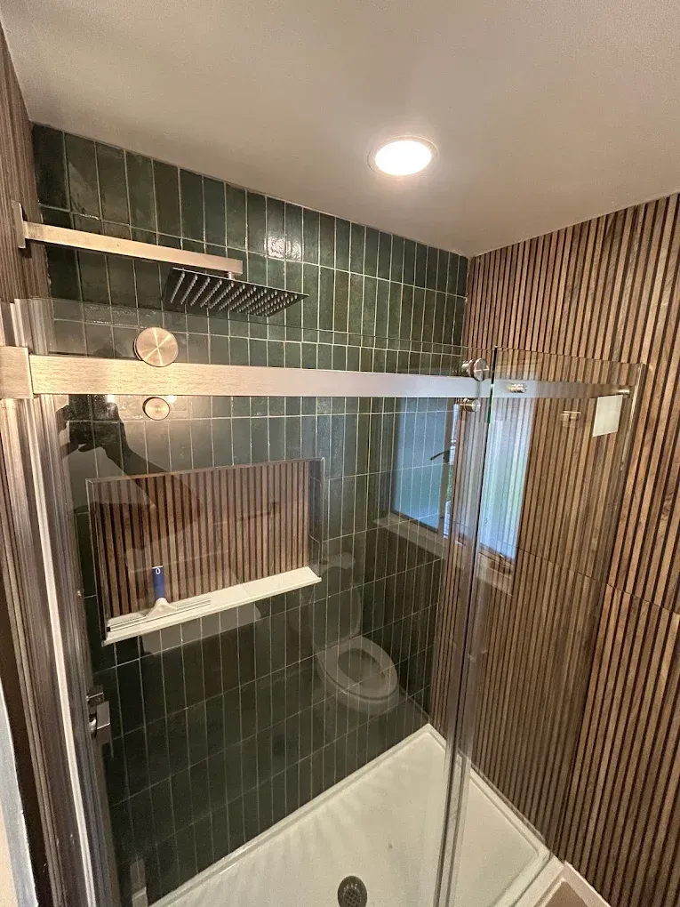 Modern bathroom with green tiled shower, wooden accent walls, glass sliding door, and overhead shower head.