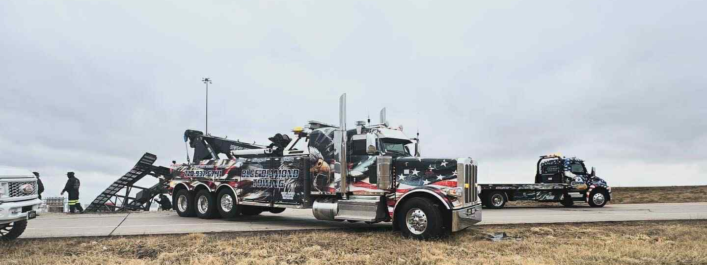 Tow trucks on a road under a cloudy sky. One large truck is hoisting wreckage. Another flatbed is nearby.
