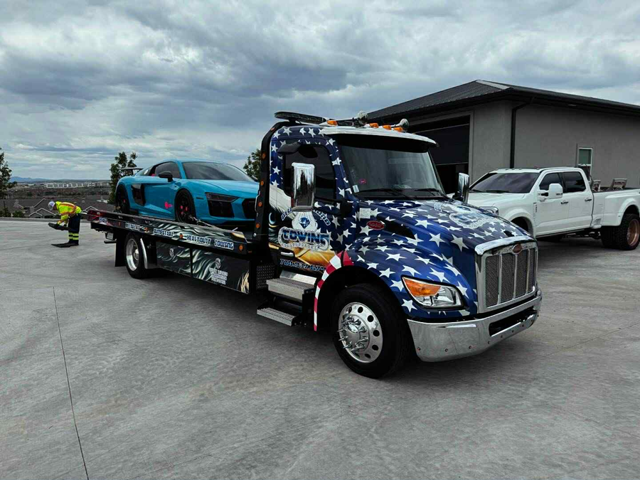 Tow truck with blue patriotic stars and stripes carrying a blue sports car, parked on concrete.