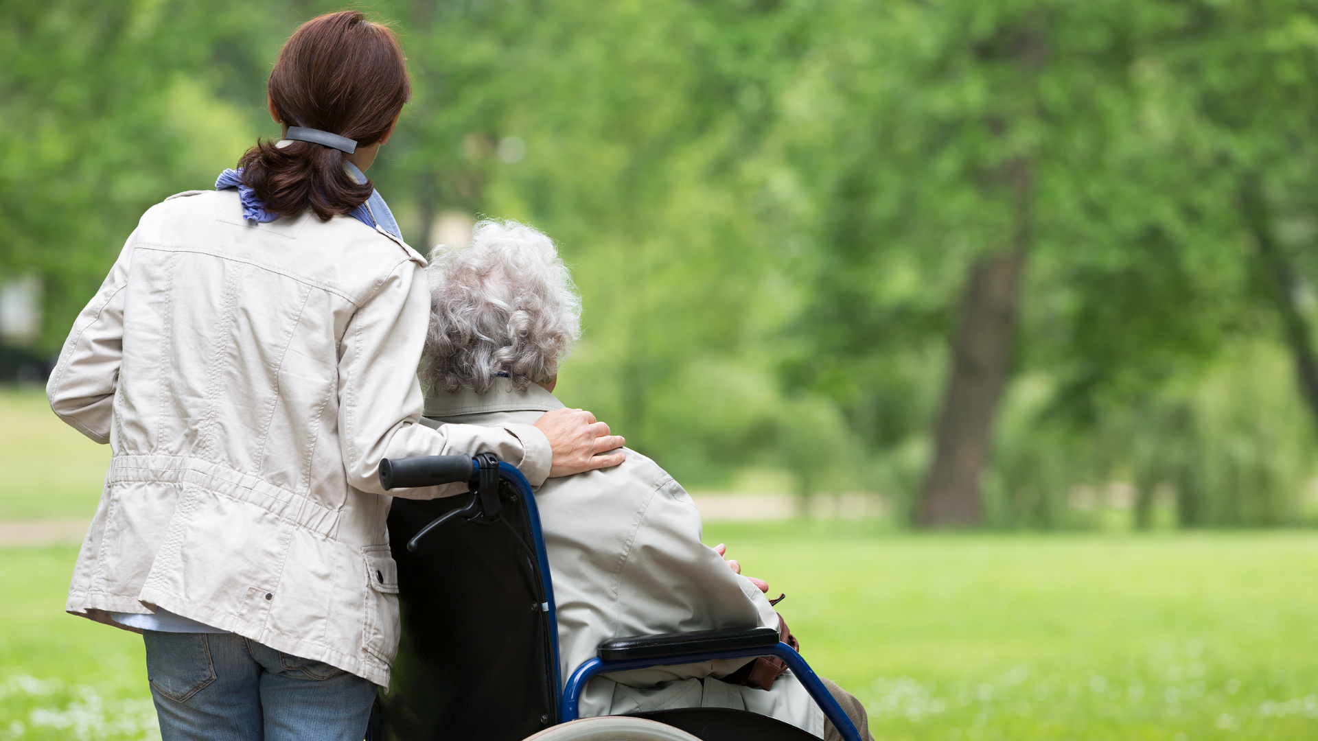 A woman is walking with an elderly woman in a wheelchair in a park.