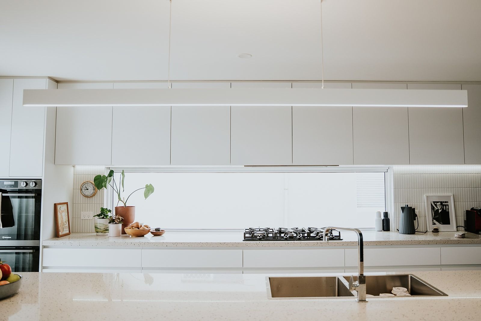 Modern Minimalist Kitchen With a Long, White Overhead Light Fixture, White Cabinetry, and a Beige Speckled Countertop — Darko's Kitchens in Kiama, NSW