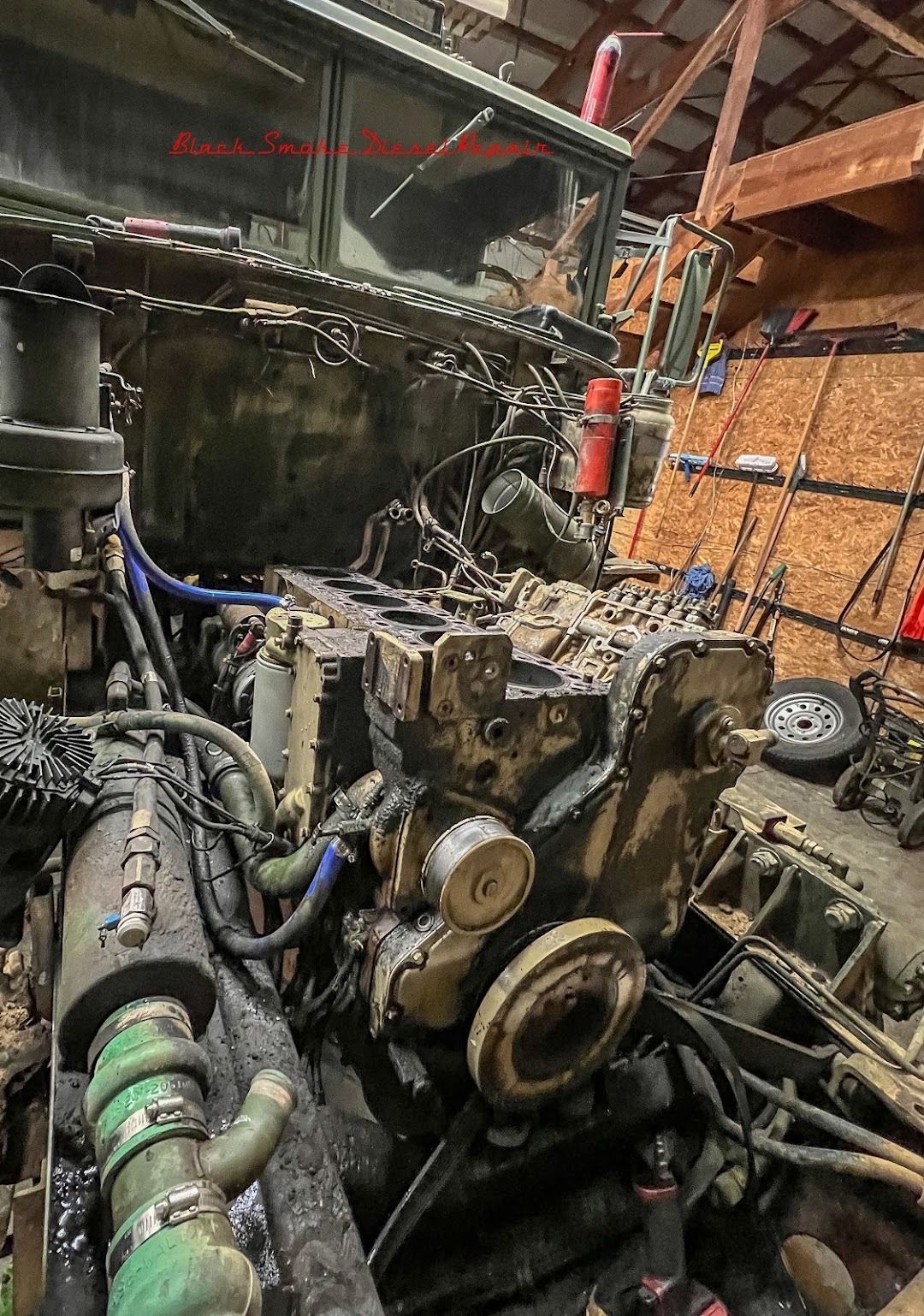 An open, dark-colored truck engine bay under repair inside a garage with wooden walls.