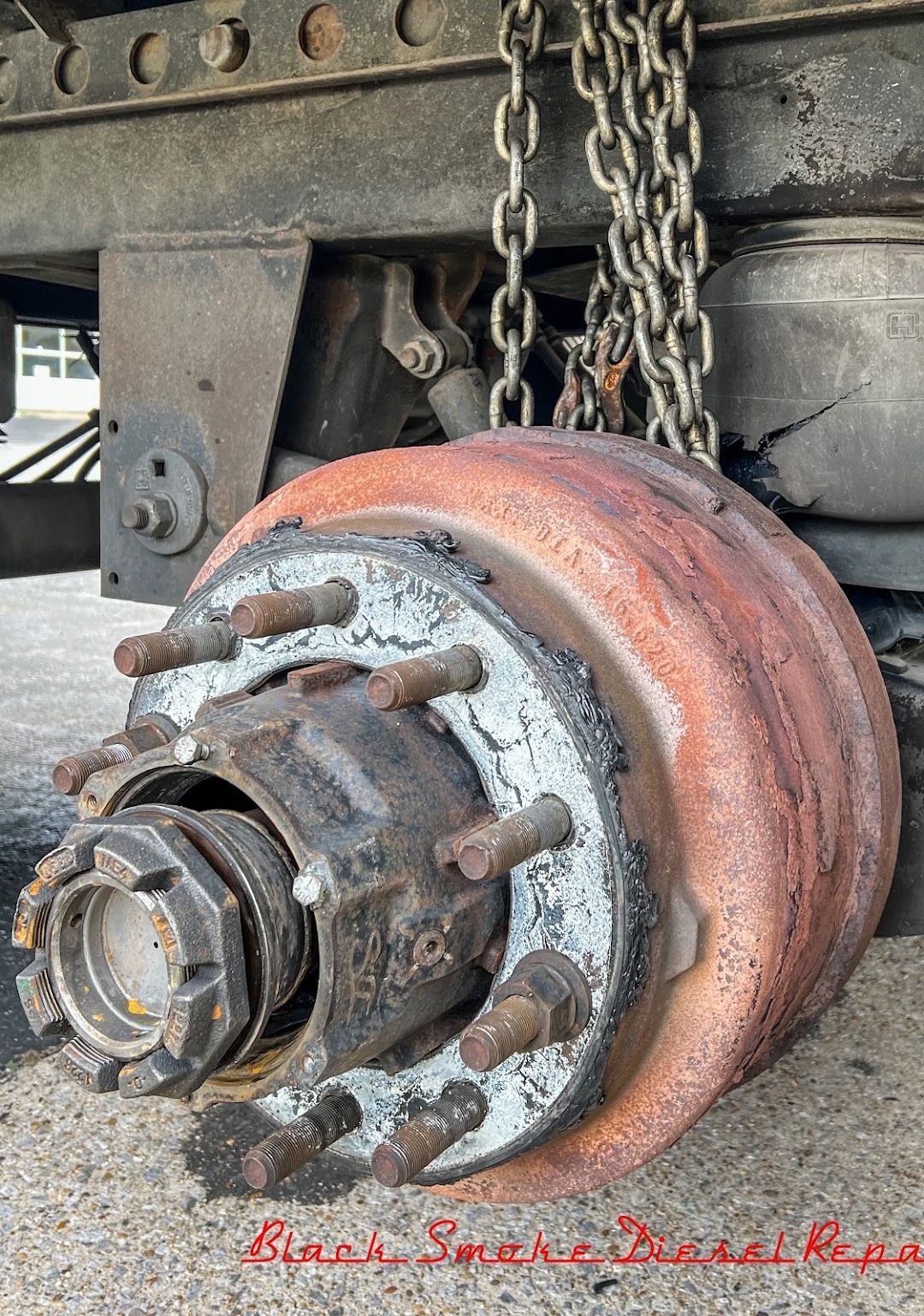 A rusty, heavy-duty truck wheel hub assembly and brake drum suspended by a chain under a vehicle frame.