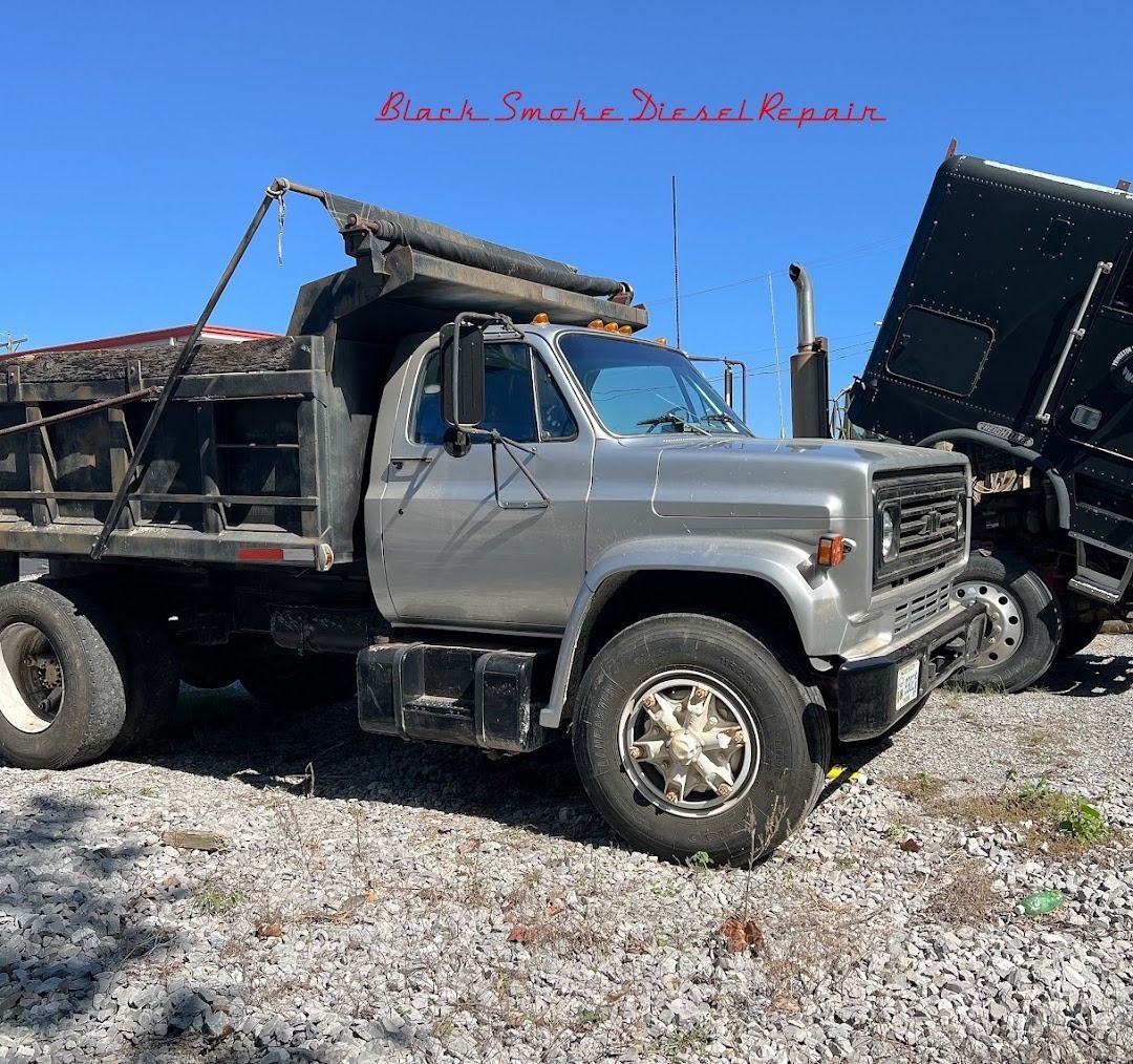 A silver dump truck parked on a gravel lot next to another large, dark truck under a clear blue sky.