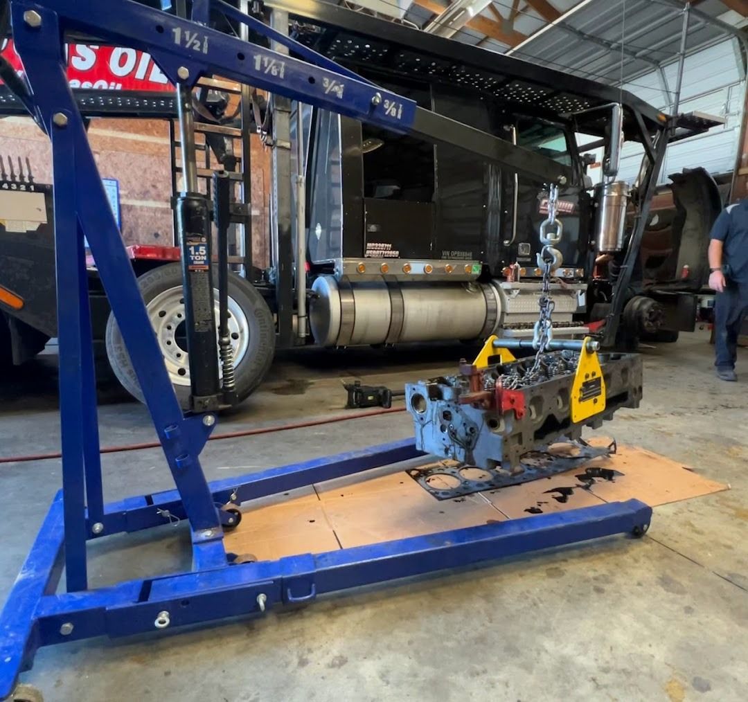 A blue engine hoist suspends a grey engine block over a cardboard mat in a garage with a large truck in the background.