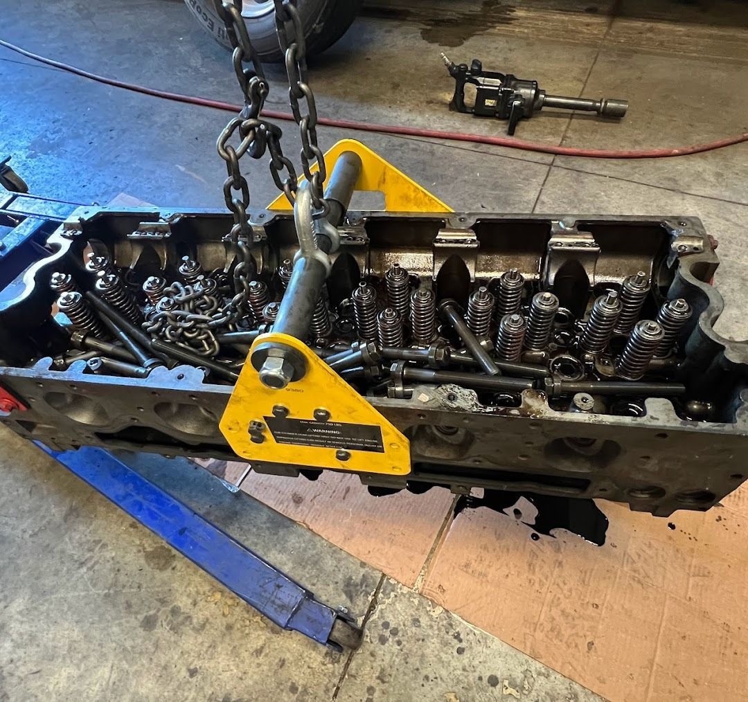 A large metal engine cylinder head suspended by a yellow lifting fixture and chains in a workshop.