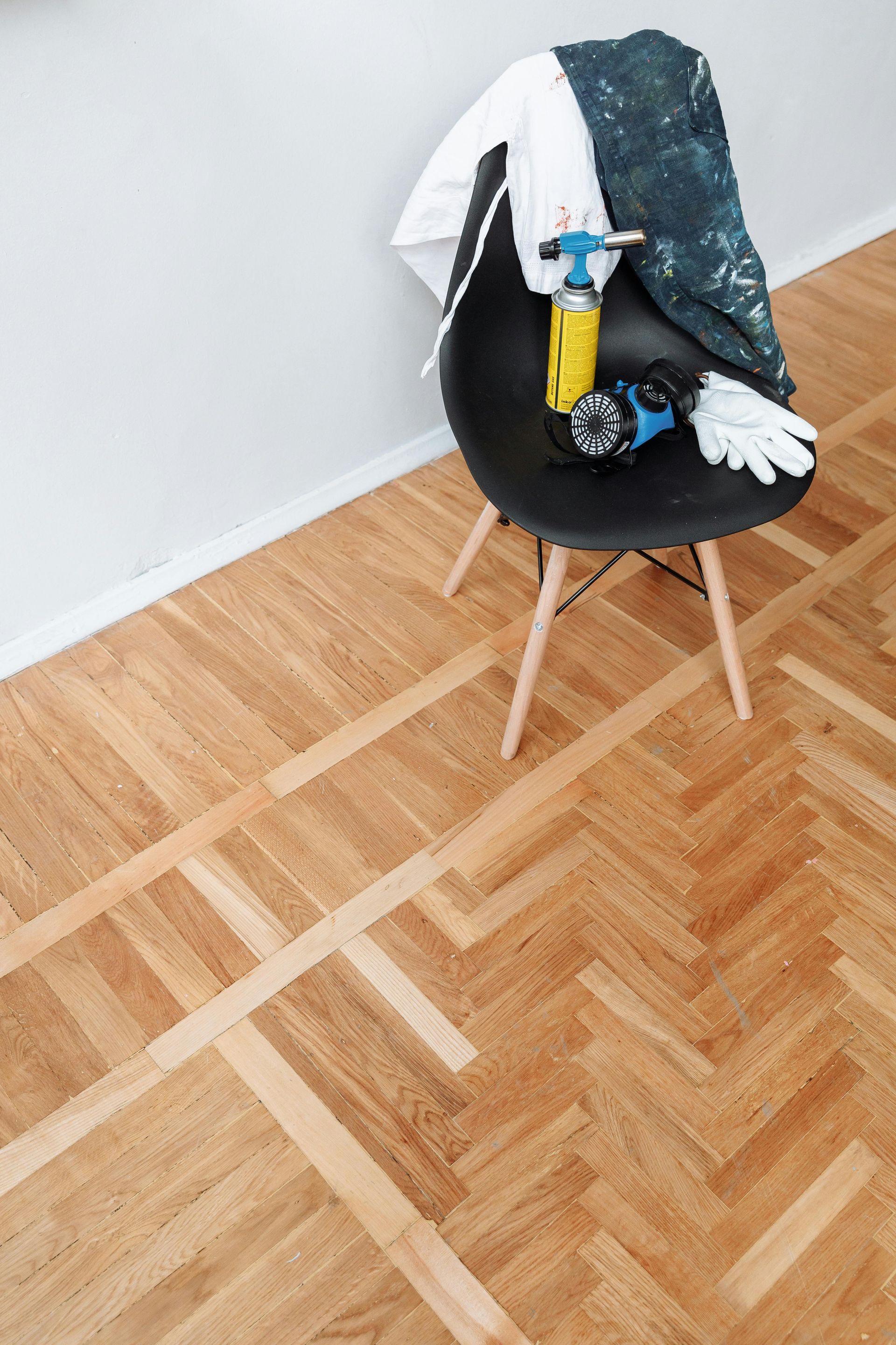 A chair with cleaning supplies and gloves, on a herringbone wood floor with taped sections for refinishing