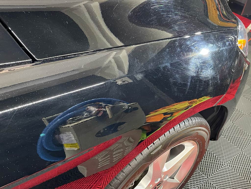 Black car's quarter panel being worked on with detailing tools, reflection of equipment visible, red detailing tape.