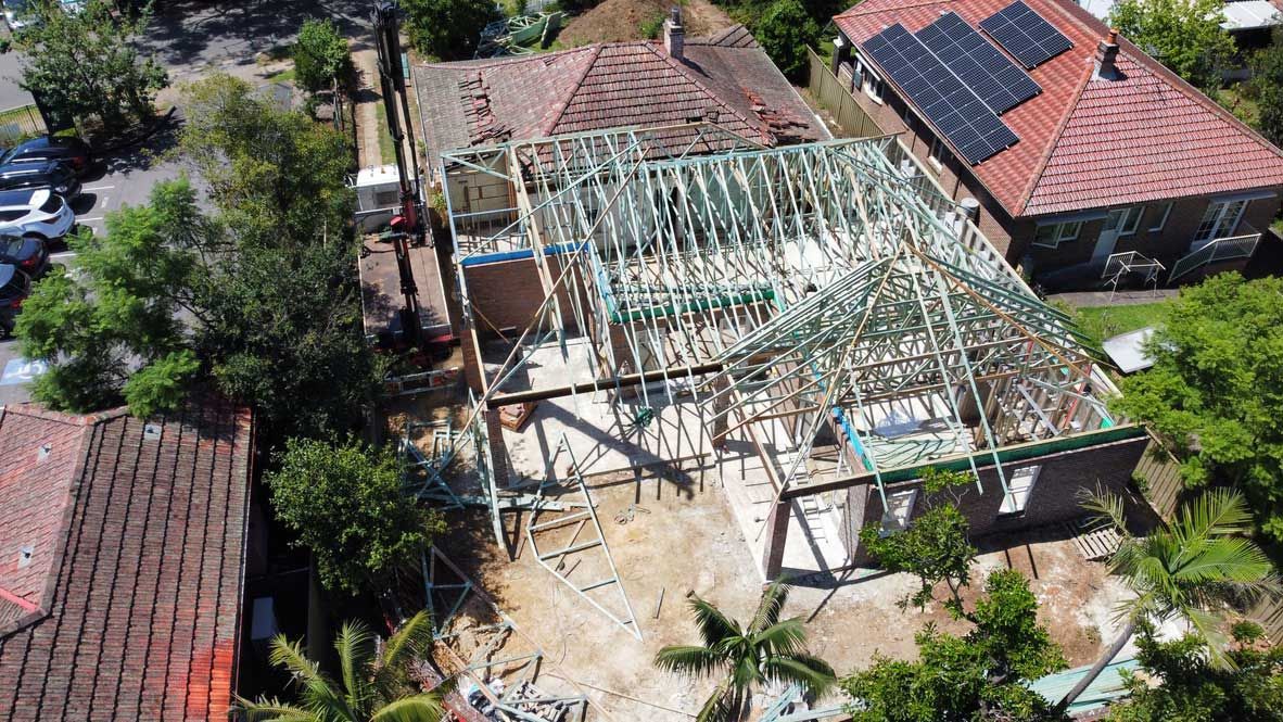 Aerial view of a house under construction beside red-roofed homes and trees, with exposed roof framing.