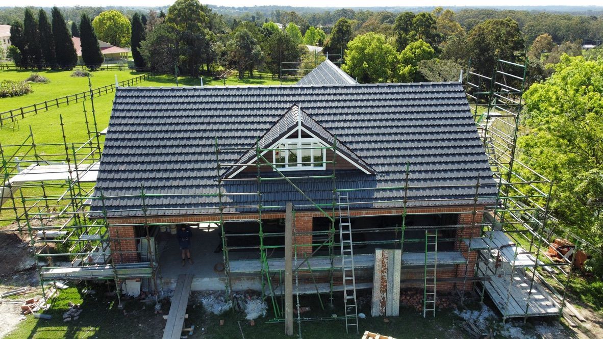 Two-story house under construction with dark gray roof, scaffolding, and green countryside in the background