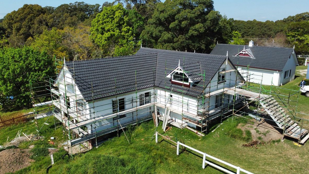 Aerial view of a house roof under construction with exposed wood framing and scattered materials surrounded by trees.