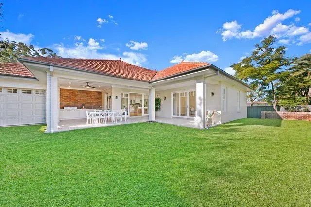 Single-story cream house with red-tiled roof, front porch, and large green lawn under a blue sky