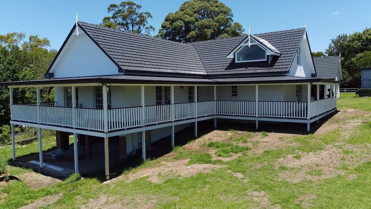 White house with wraparound porch and dark tiled roof on a grassy, sunny hillside