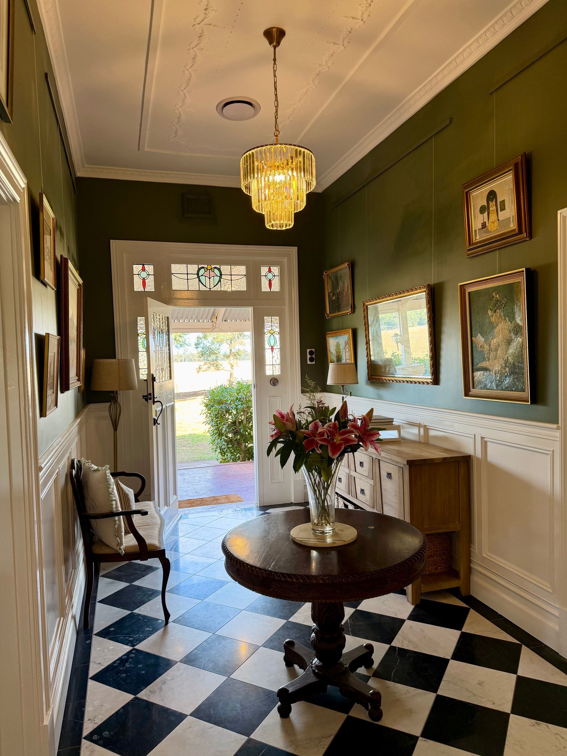 Narrow hallway with black-and-white checkered floor, green walls, chandelier, framed art, and small table with flowers