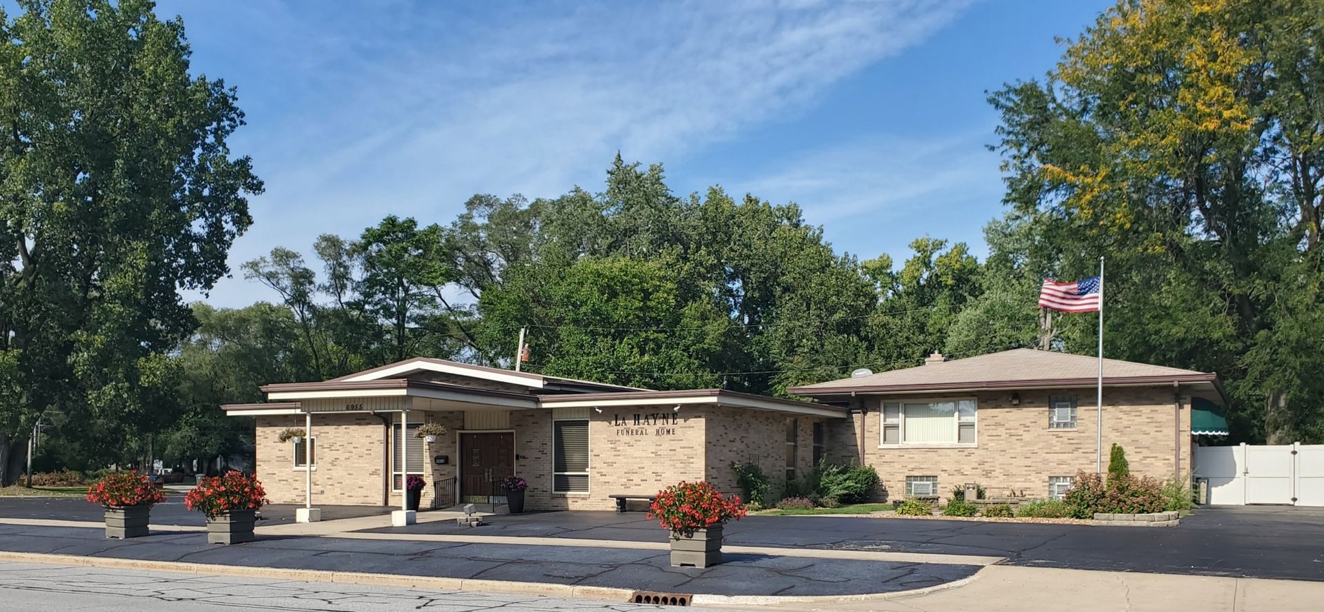 A one-story brick building with trees in the background under a blue sky. There are flower pots in front.
