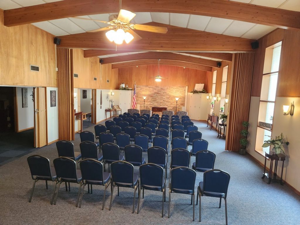 Rows of blue chairs in a wood-paneled funeral home chapel. American flag hangs near the front.