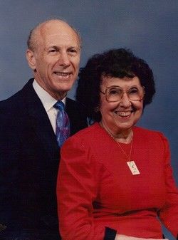 Man and woman smiling, posed for portrait. Man in suit, woman in red top with glasses.