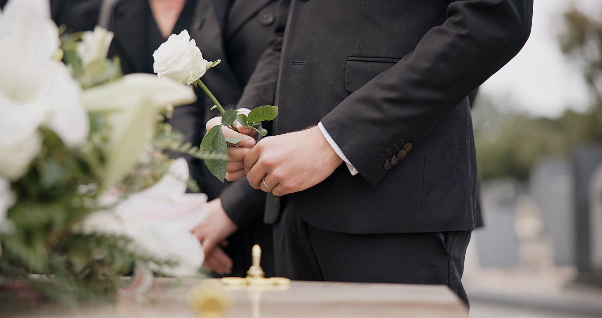 Person in black suit holding a white rose at a funeral, near a casket and flowers.