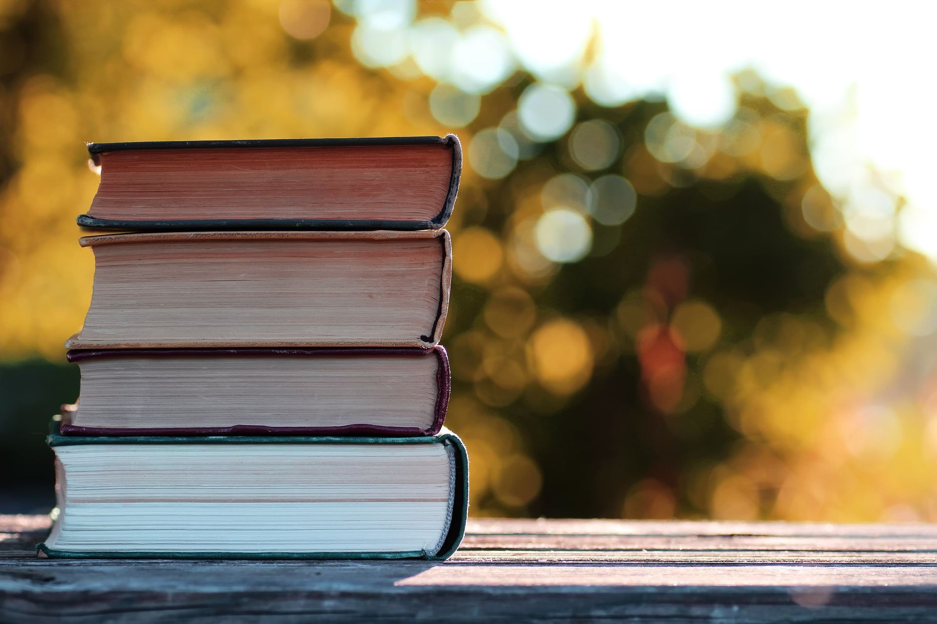 Stack of old books on a wooden surface, with blurred autumn foliage in the background.