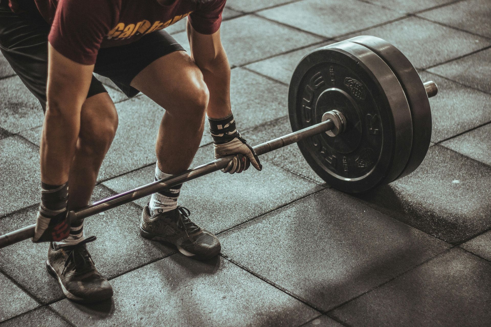 Man lifting a barbell in a gym; wearing gloves, shorts, and athletic shoes, on a rubber floor.