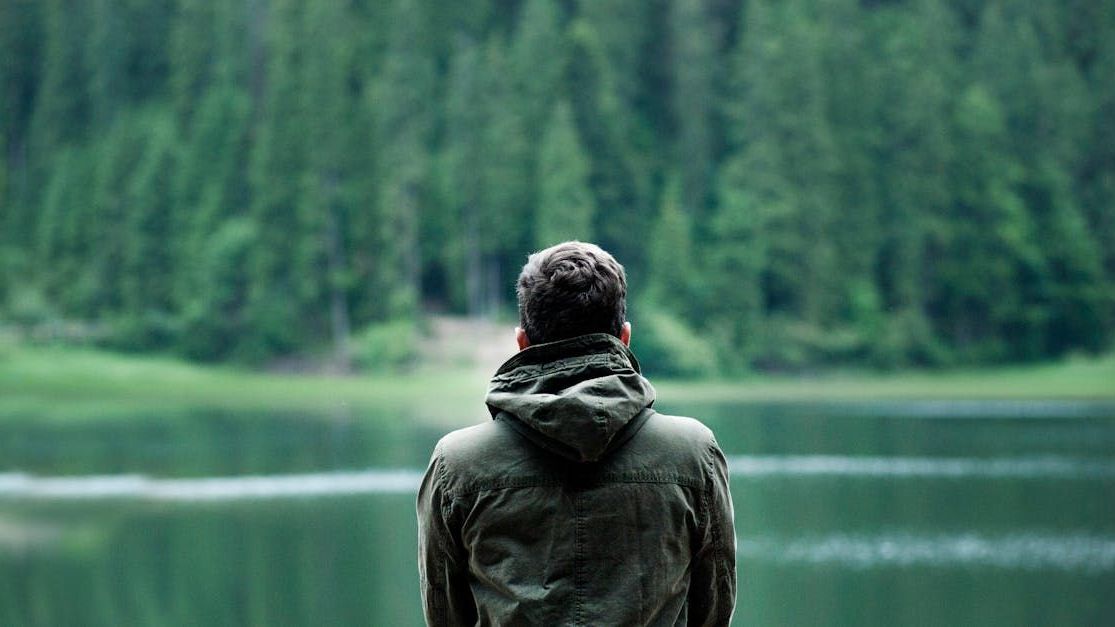 A man is standing in front of a lake with trees in the background.