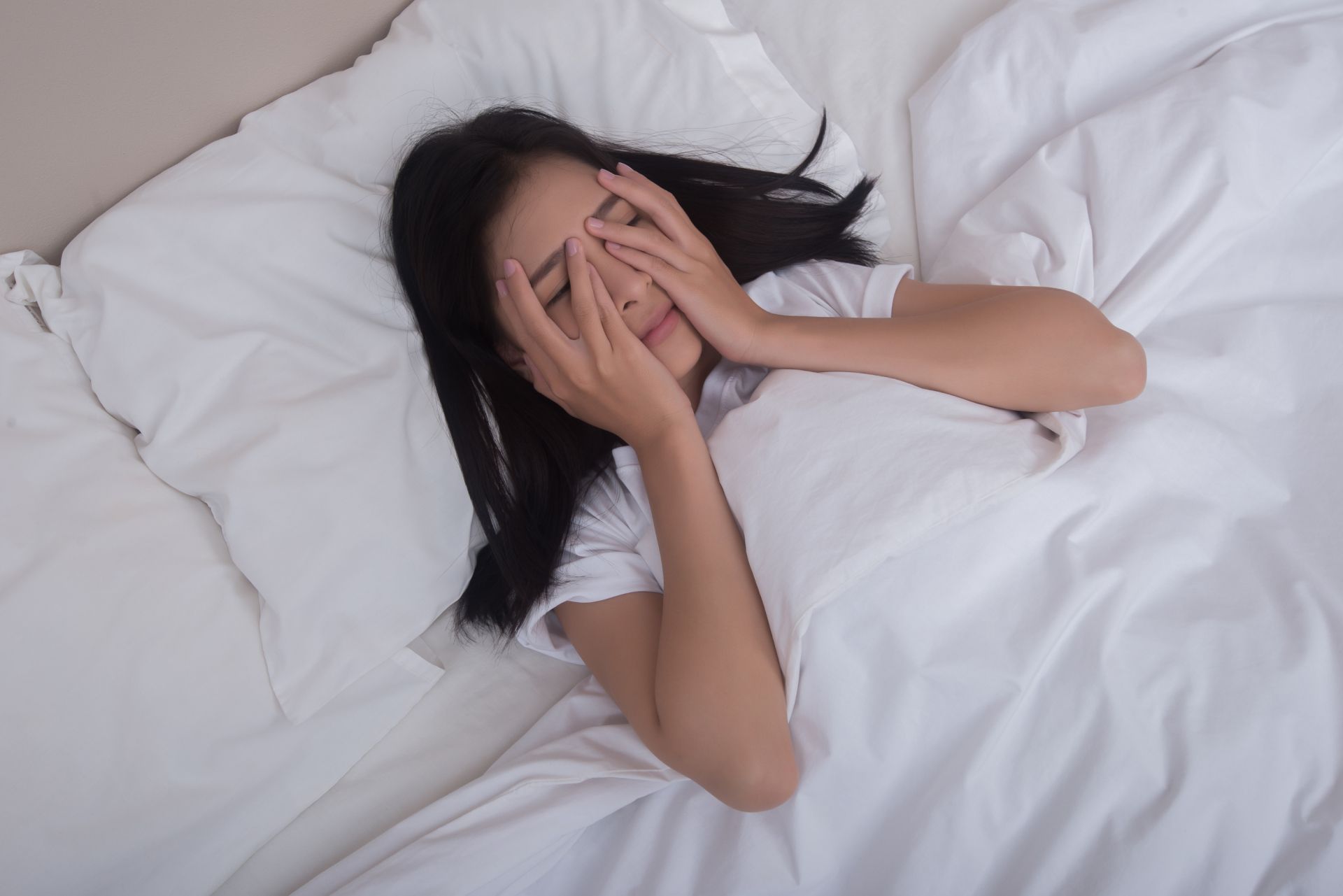 A woman is laying in bed covering her face with her hands.