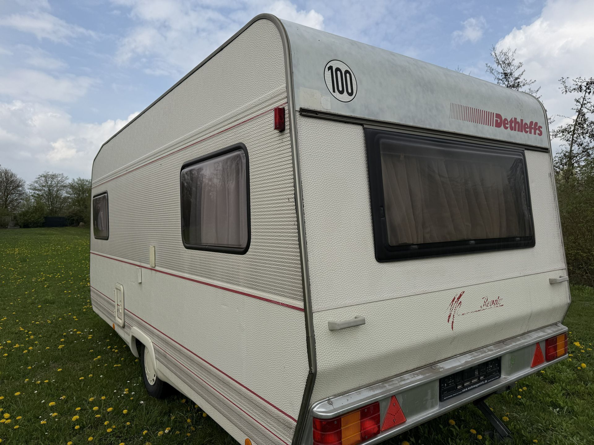 Ein cremefarbener Dethleffs-Wohnwagen mit einem „100“-Geschwindigkeitsaufkleber, geparkt auf einer grasbewachsenen Wiese unter bewölktem Himmel.