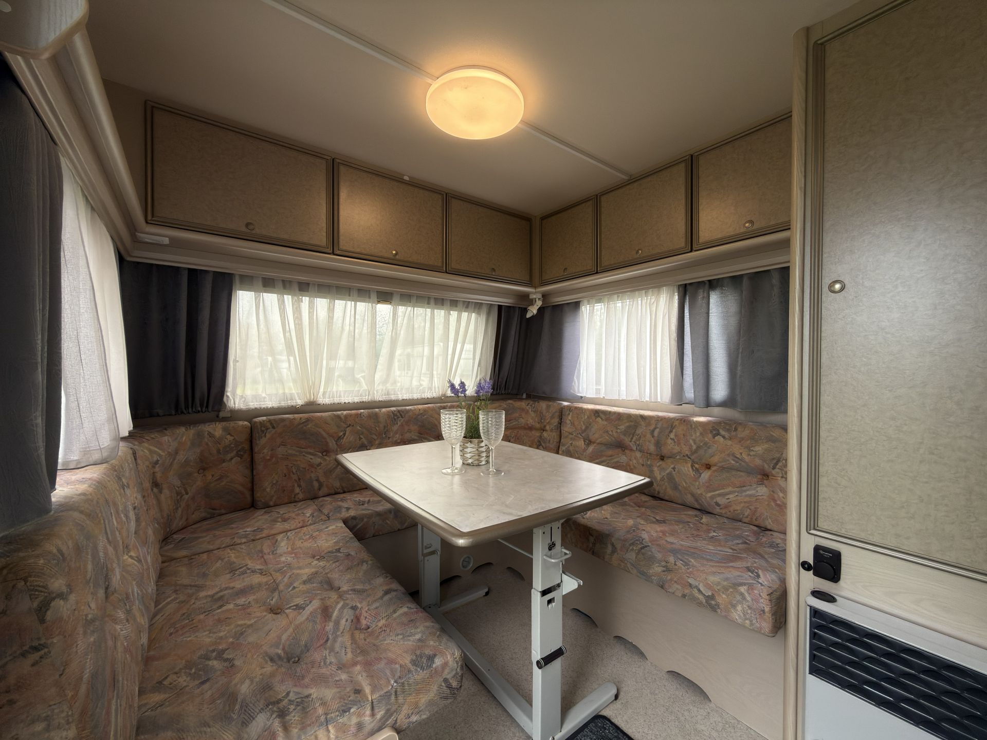 Innenansicht eines Wohnmobils mit einer U-förmigen Sitzbank, einem zentralen Tisch, hellen Holzschränken und Vorhängen.