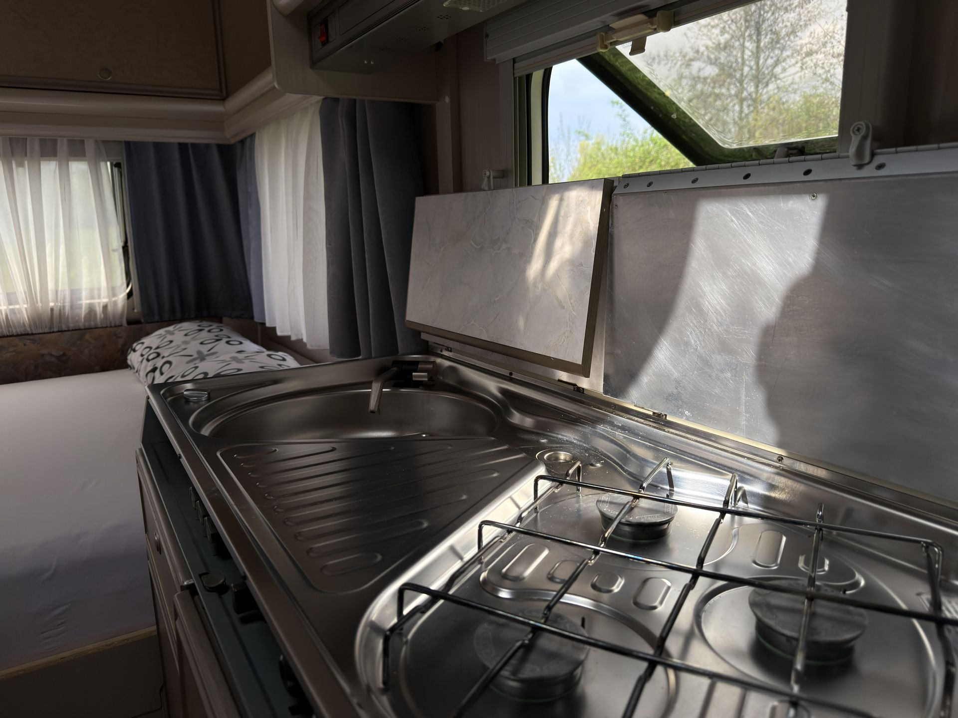 In einem Wohnmobil befinden sich eine Spüle aus Edelstahl und ein Gaskochfeld neben einem Bett mit einer gemusterten Decke.