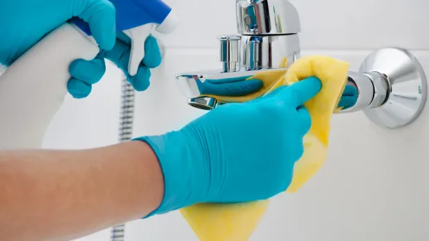 Person wearing blue gloves cleans a chrome faucet with a yellow sponge and spray bottle in a white bathroom.