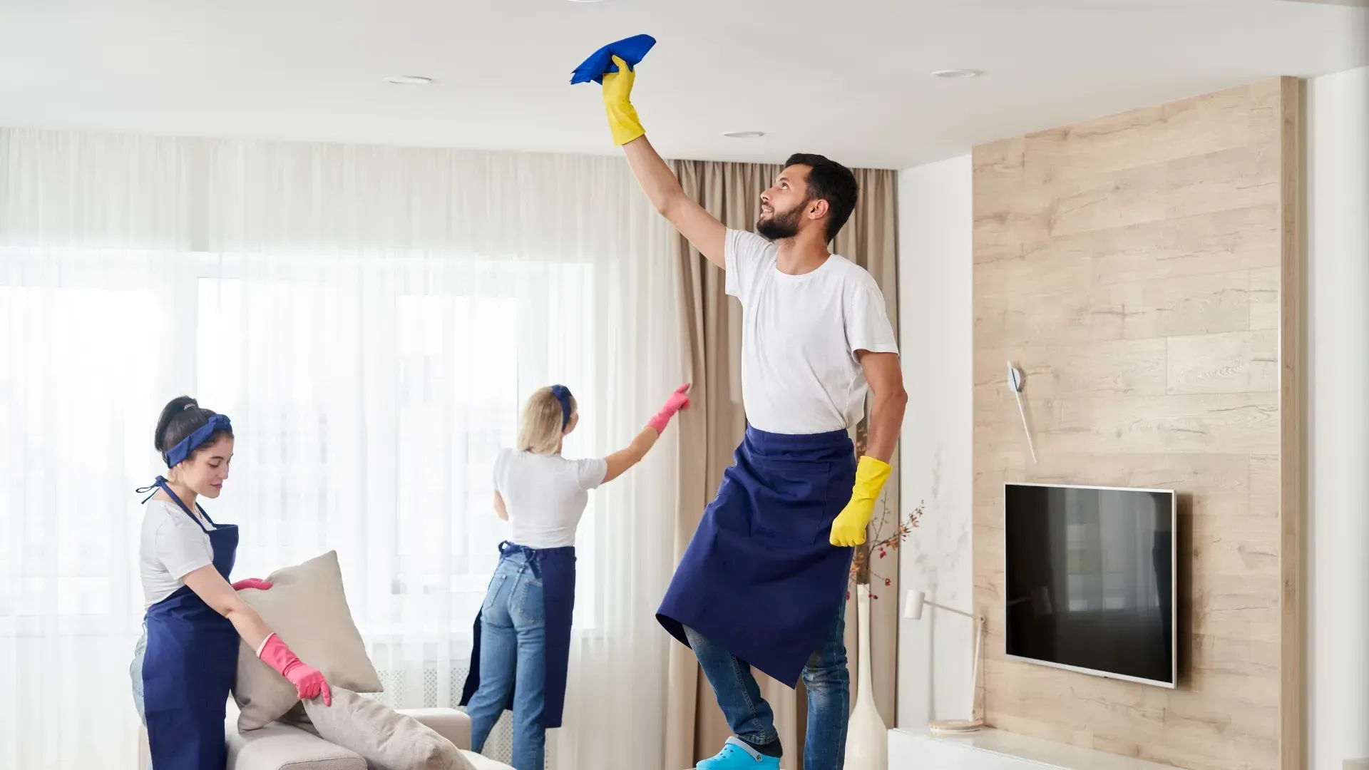 Three people cleaning a bright living room: One on a ladder cleaning the ceiling, two others tidying furniture.