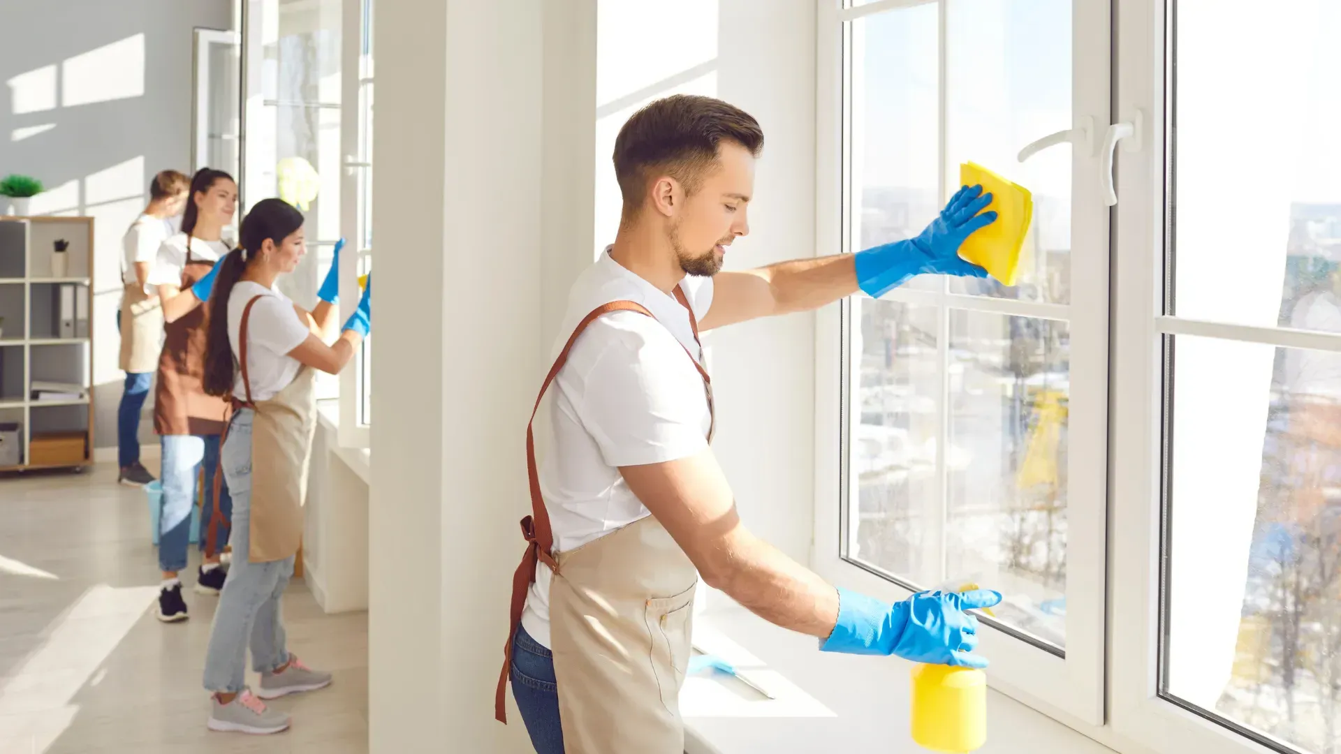 Cleaning crew wiping windows; man sprays cleaner, wearing apron and blue gloves. Other workers in background.