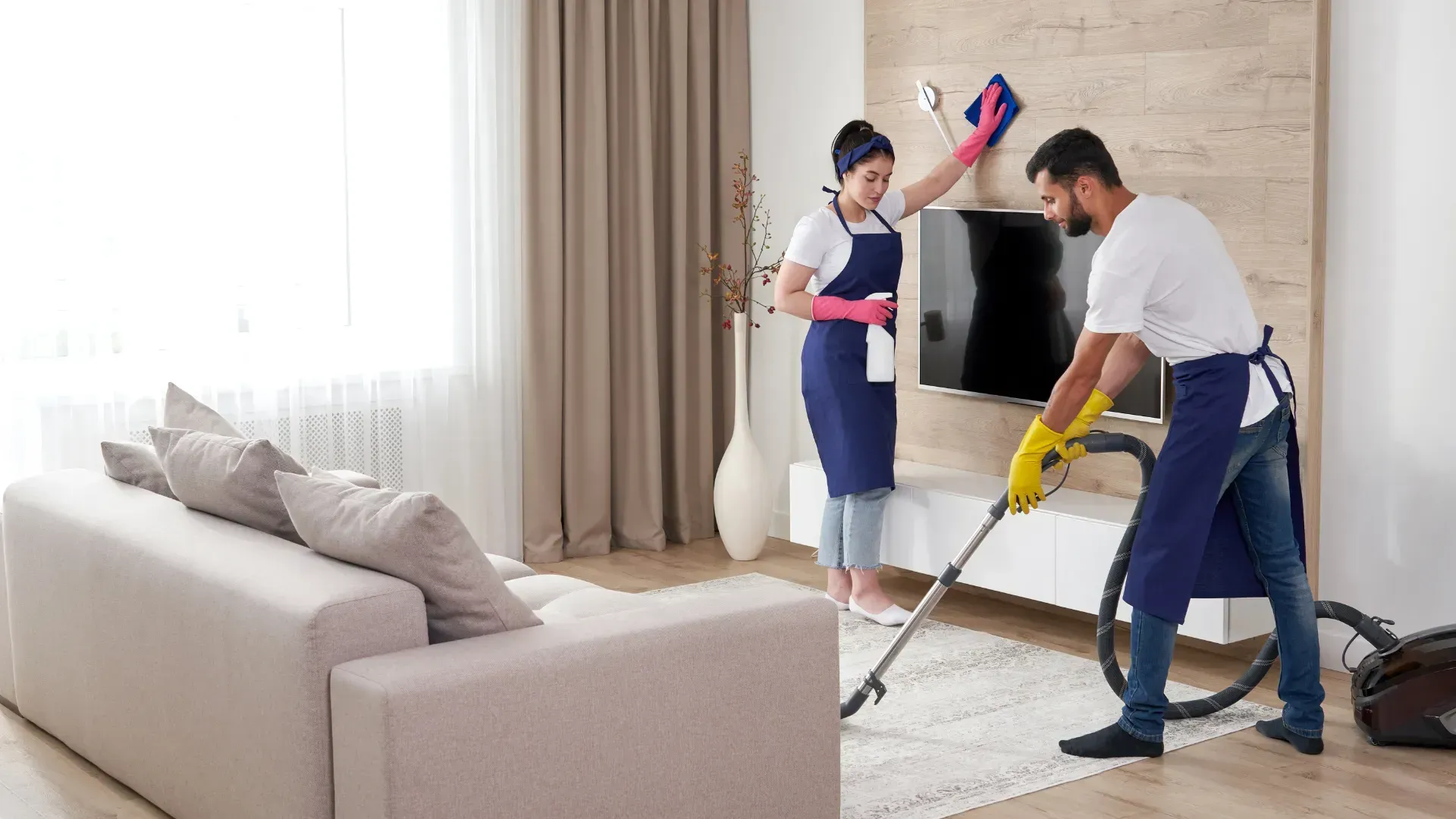 Three people cleaning a living room: one vacuuming, two dusting a wall, near a sofa and window.